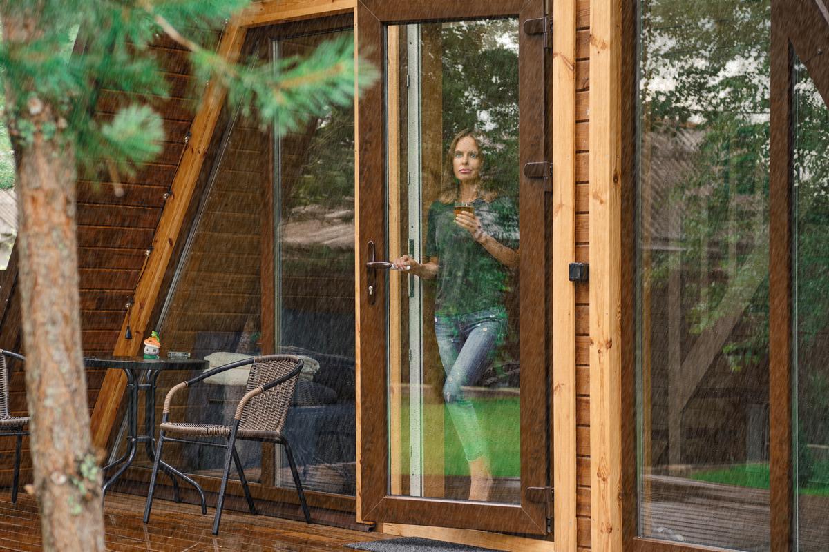 Young woman with a cup of herbal tea stands on the doorstep of a forest house and looks at the rain